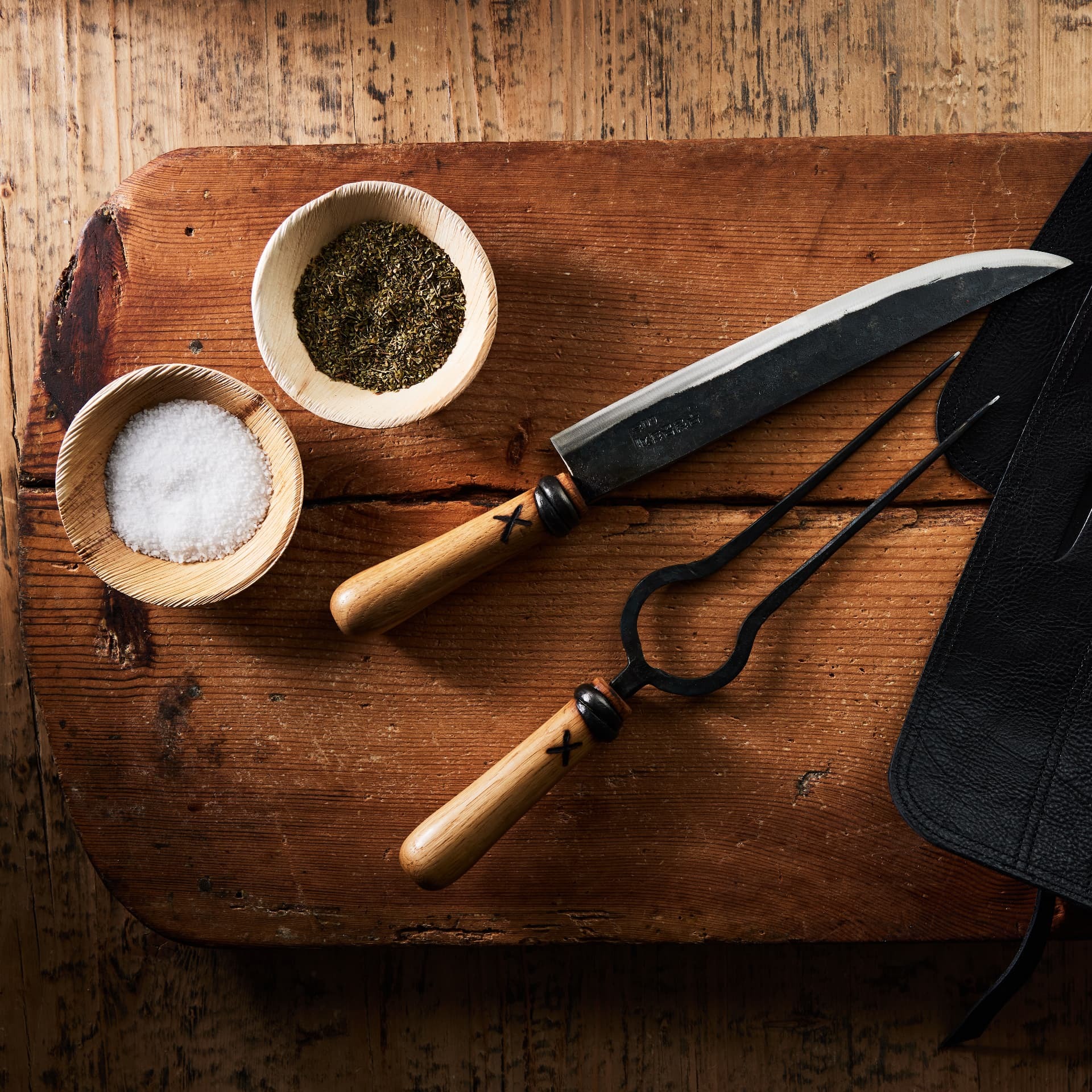 Forged knife and carving fork beside bowls of salt and herbs on a wood board
