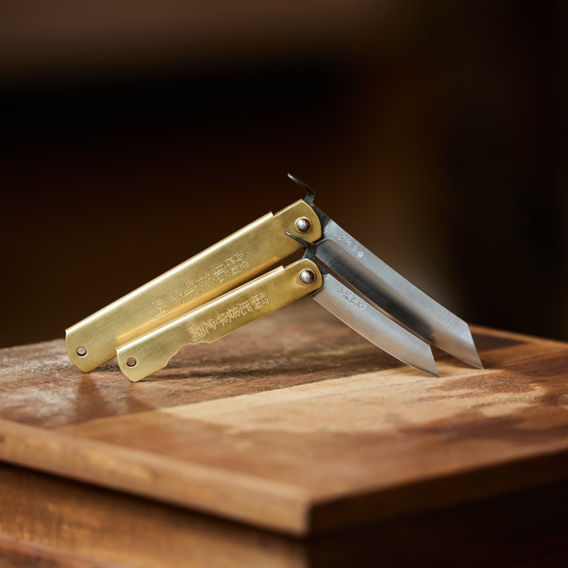 Two brass-handled Japanese folding pocket knives open on a wooden board