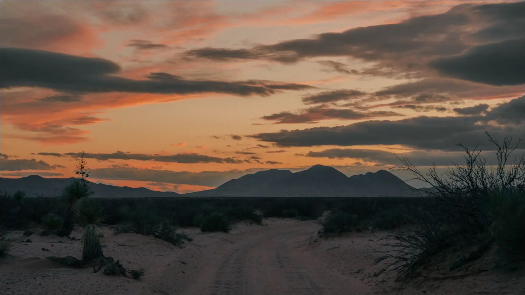 Sand two-track leading toward desert mountains under an orange sunset sky