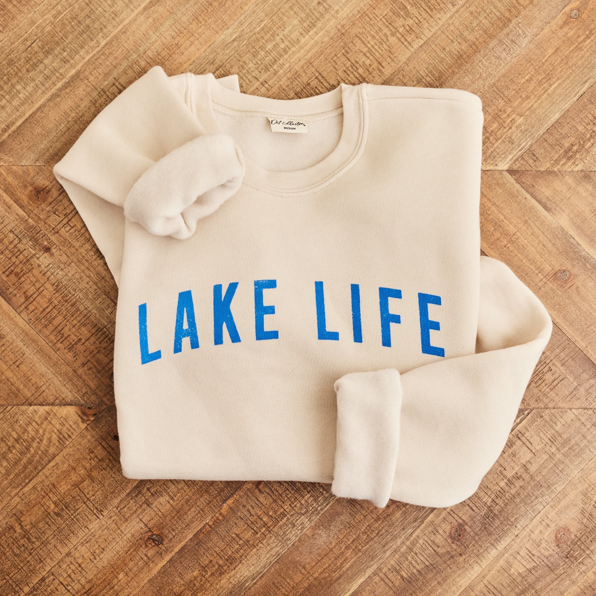 Folded cream sweatshirt with blue Lake Life lettering on a herringbone wood floor